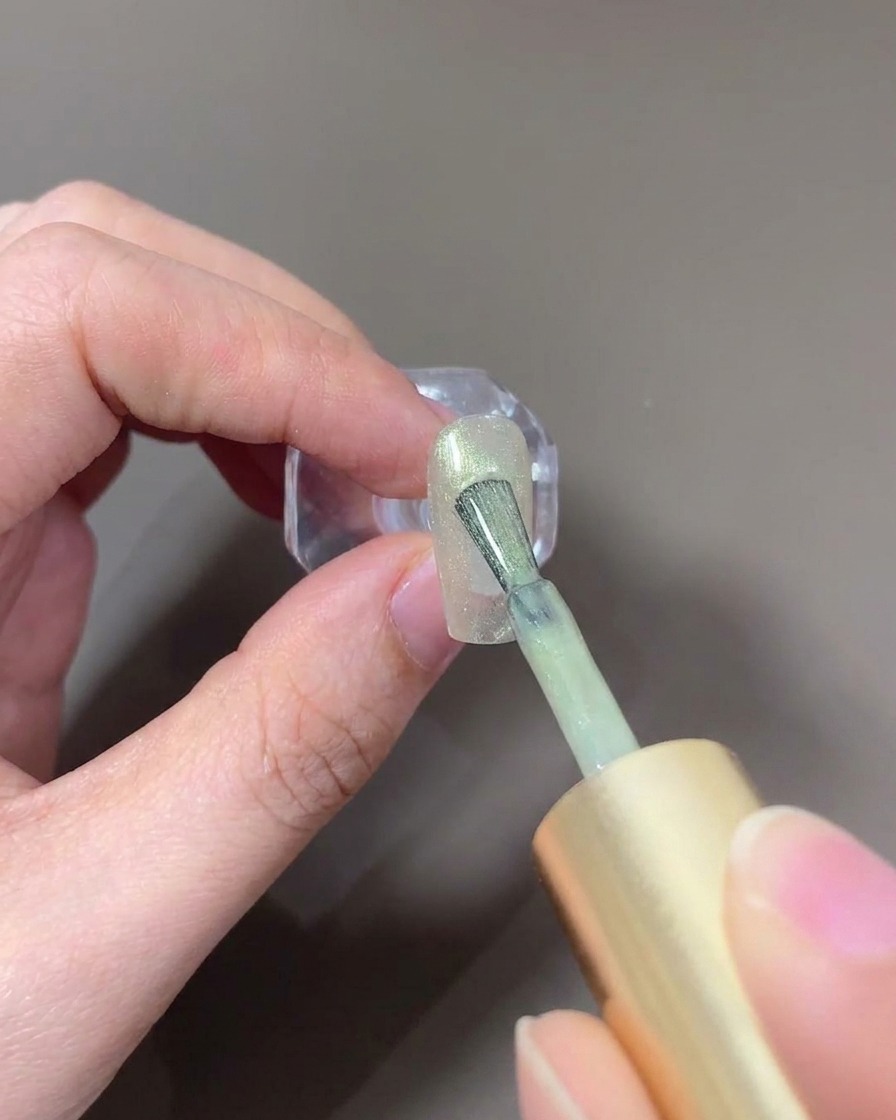 Un pinceau appliquant un gel cat-eye clair effet verre sur un ongle.