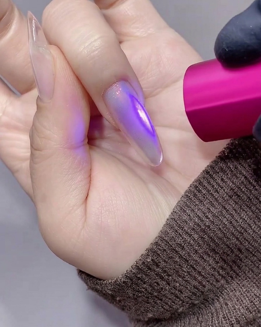 Gros plan dune main avec un ongle stiletto violet translucide catalysé sous une lampe UV/LED rose.