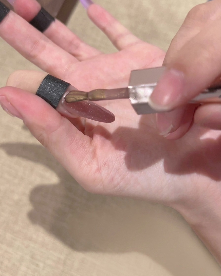 Pinceau dun flacon de vernis gel appliquant une couche de gel magnétique doré cat-eye sur un ongle avec un dégradé violet déjà présent.