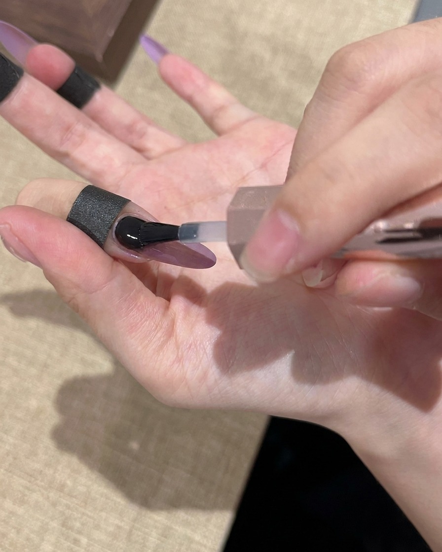 Un pinceau appliquant une finition transparente et brillante sur un ongle en gel cat-eye violet et doré.