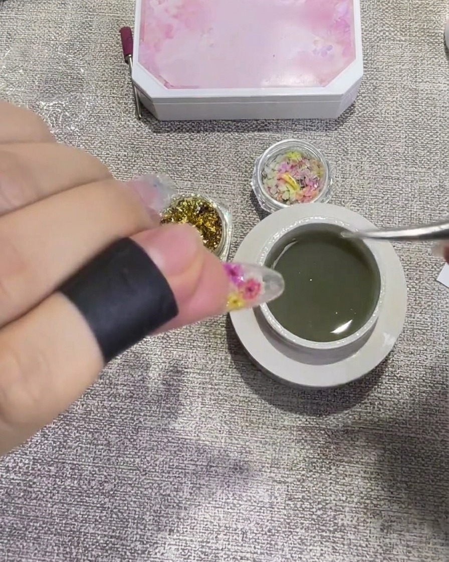 Gros plan dune pince pressant un petit morceau de feuille dor sur un ongle recouvert de gel transparent.
