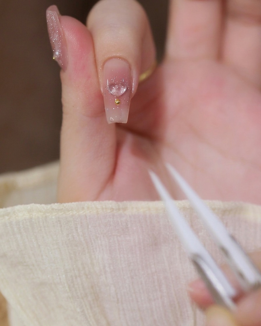 Gros plan dun index avec une base nude laiteuse, une breloque cœur en 3D et une perle dorée.