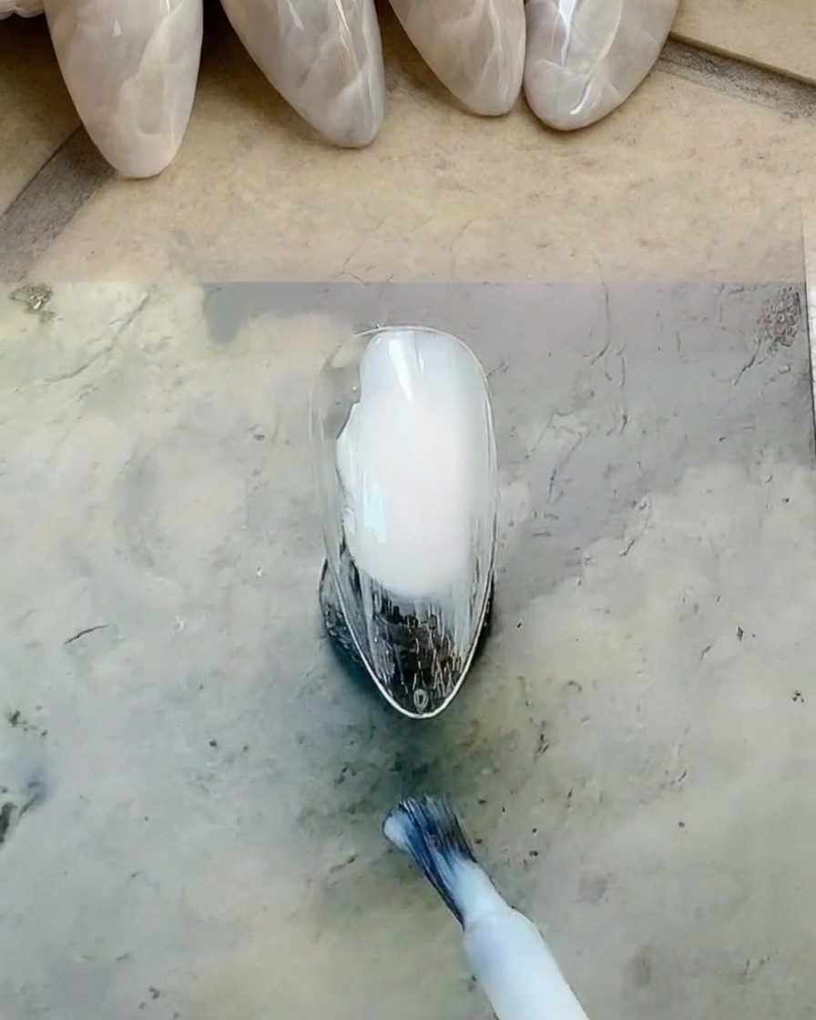 Pinceau appliquant un gel blanc laiteux sur la surface de longle.