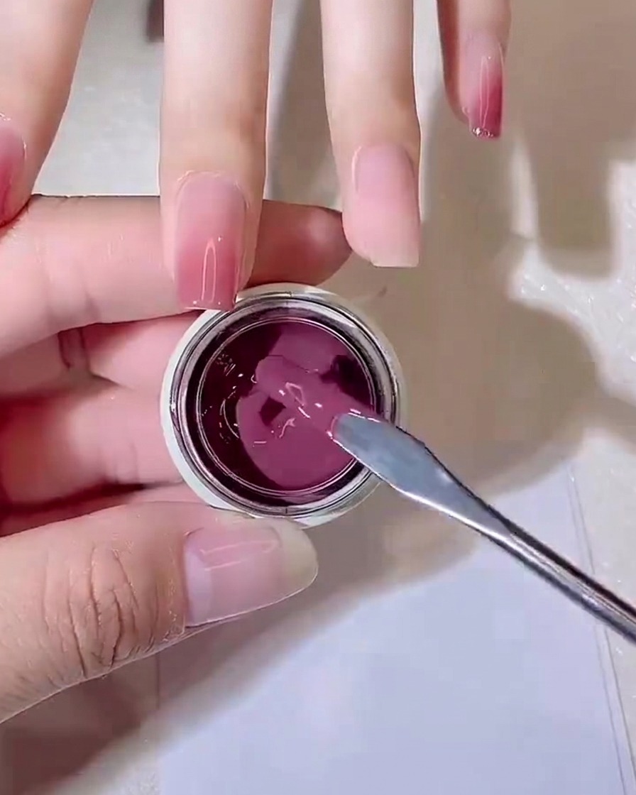 Un bâtonnet mélangeur en métal prélevant une petite noix de gel jelly rose violacé dans un petit pot.