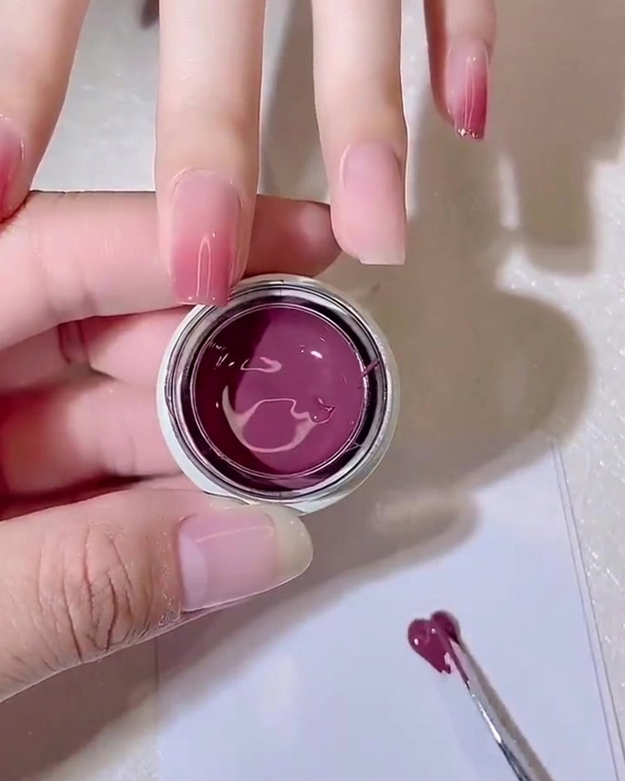Un outil en métal étalant une petite quantité de gel rose violacé en une fine couche sur une palette en papier blanche.