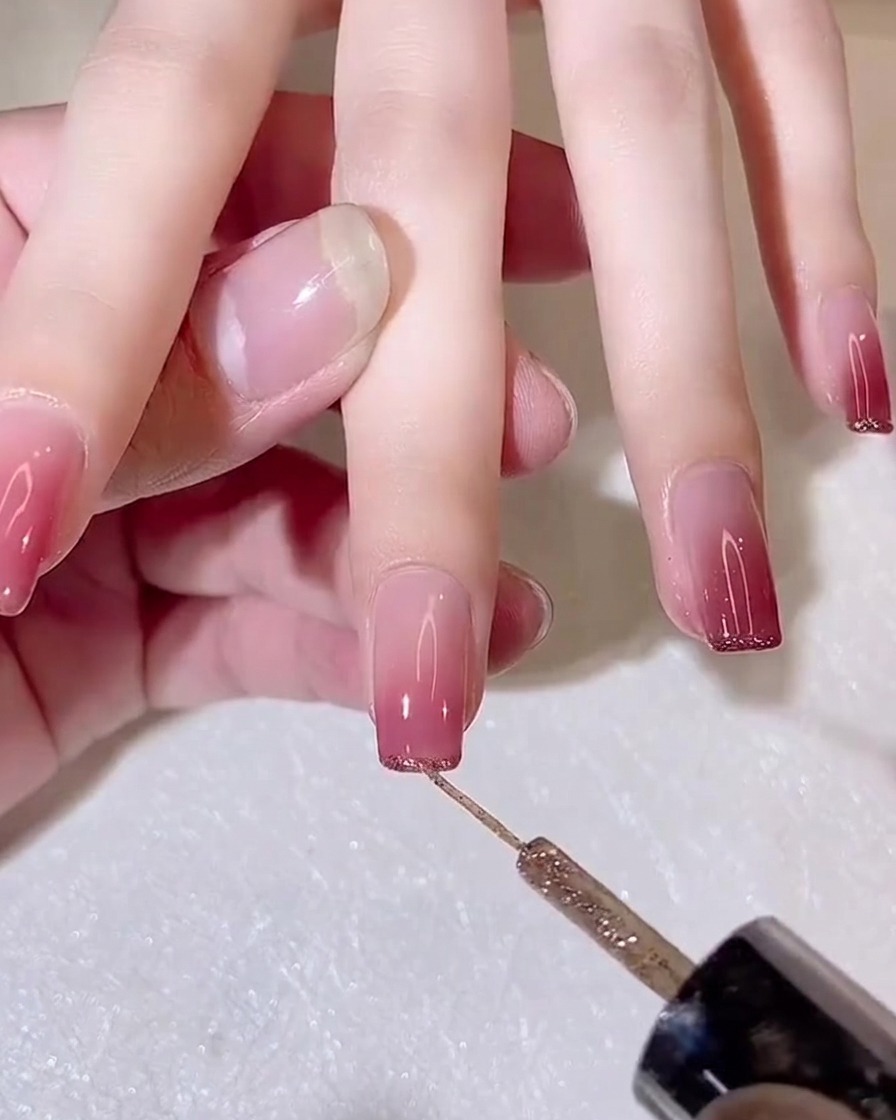 Un pinceau fin liner appliquant une fine ligne de gel pailleté doré sur le bord libre dun ongle en dégradé.