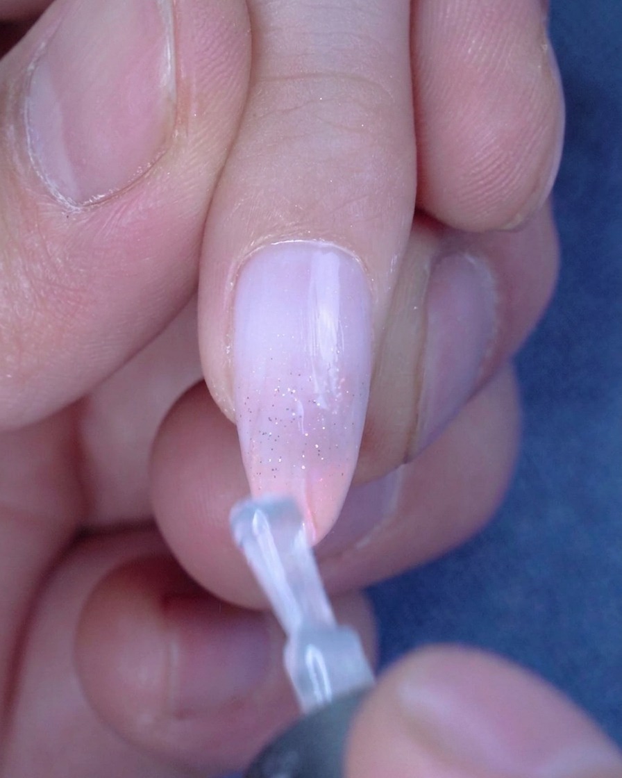 Un pinceau appliquant une couche de top coat brillant sur un ongle ombré pailleté.