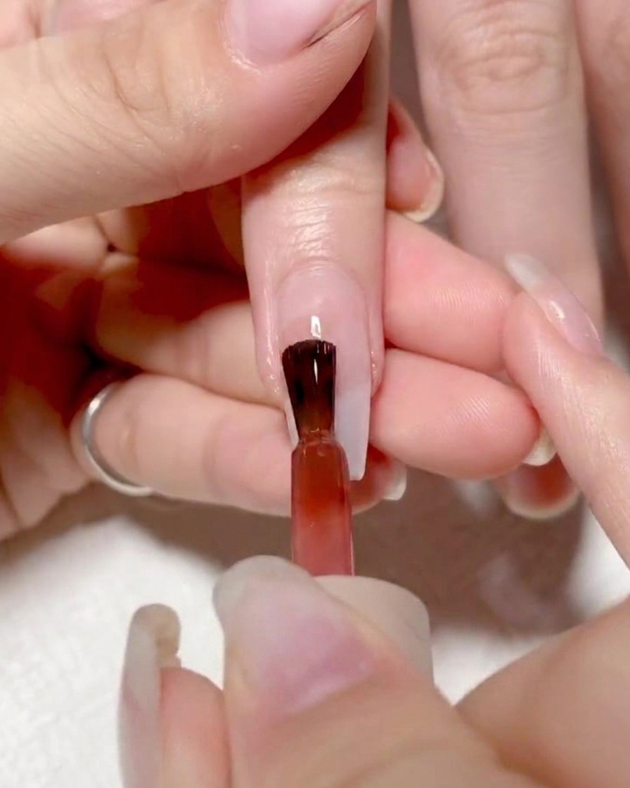 Un pinceau appliquant un gel rouge vif sur la partie gauche de longle.
