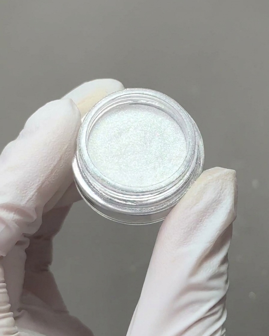 Une main gantée tenant un petit récipient de poudre chrome aurora irisée avec un embout applicateur en éponge prélevant un peu de pigment.