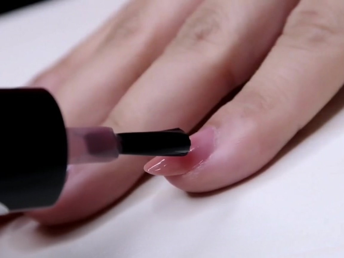 Un pinceau appliquant une couche de vernis rose transparent sur la moitié extérieure dun ongle.