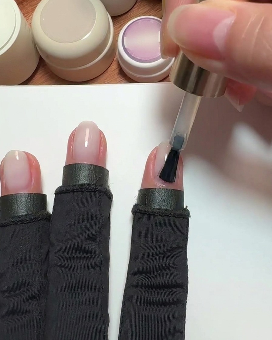 Un pinceau appliquant un vernis gel nude rosé transparent sur un ongle dindex.