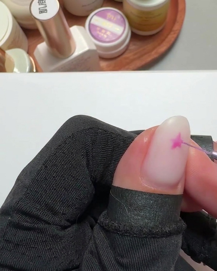 Gros plan dun pinceau fin traçant une forme détoile rose vif sur un ongle à base blanche laiteuse.