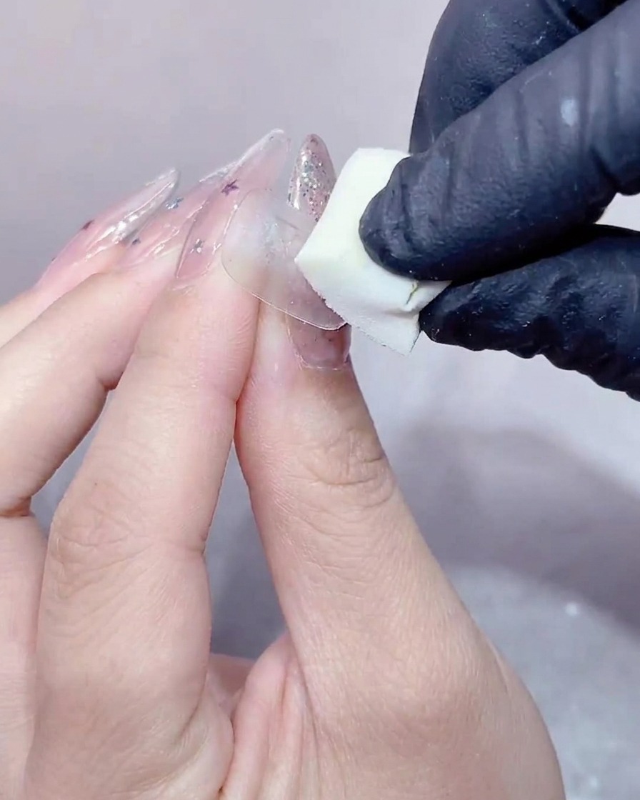 Une main gantée pressant une petite éponge à maquillage blanche sur la surface dun ongle recouvert de gel transparent.