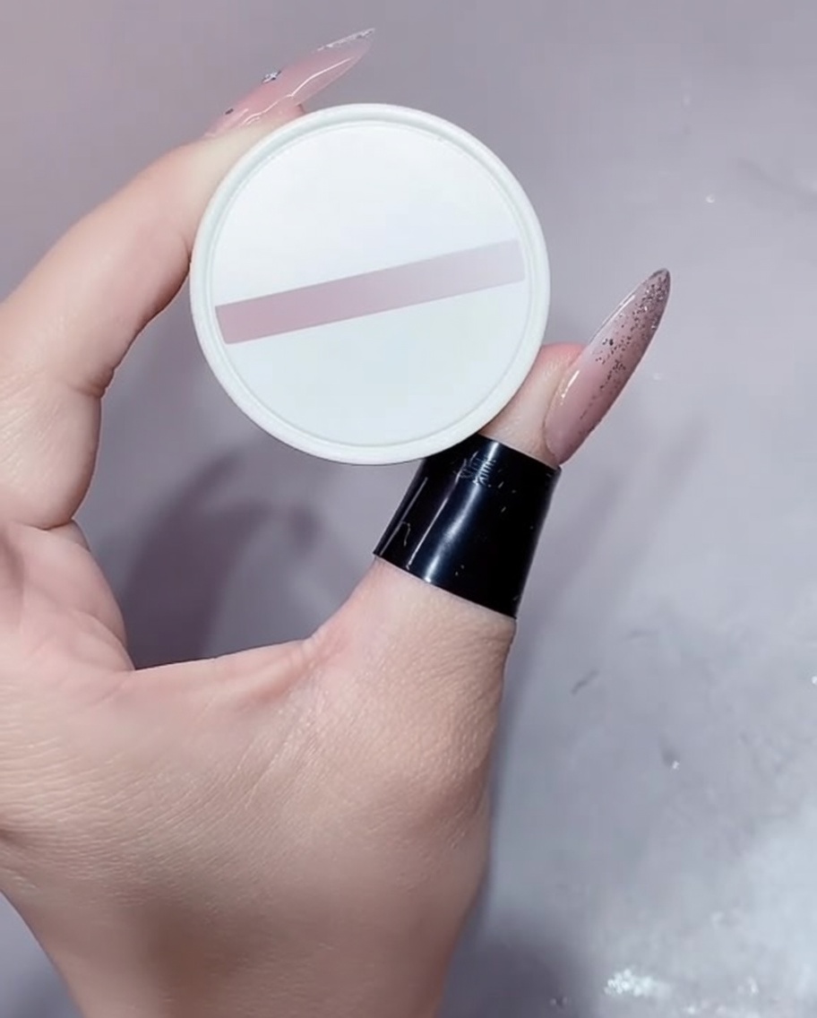 Un petit pot blanc de vernis gel nude tenu par une main manucurée.
