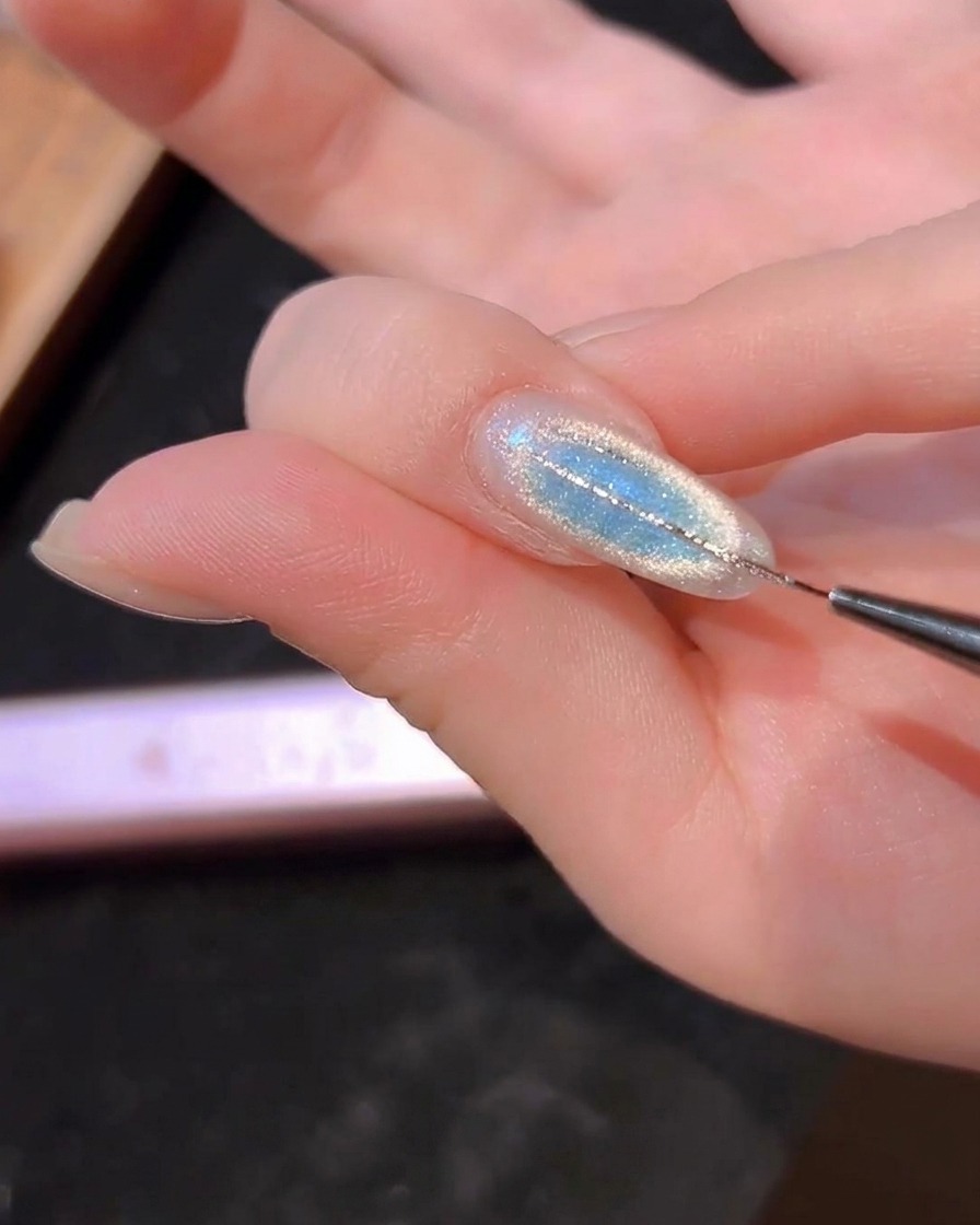 Un pinceau liner fin traçant de fines lignes argentées sur un ongle bleu œil-de-chat.