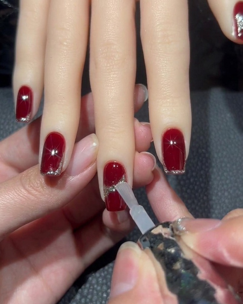 Un pinceau appliquant un top coat transparent sur un ongle rouge avec un design pailleté, en évitant soigneusement le strass central.