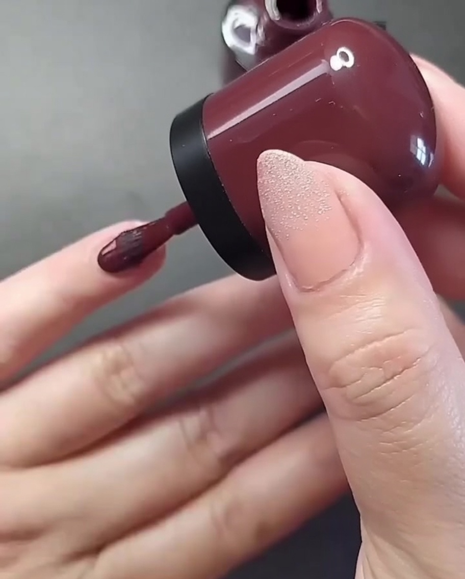 Gros plan sur une main appliquant un vernis bordeaux profond sur longle de lauriculaire.