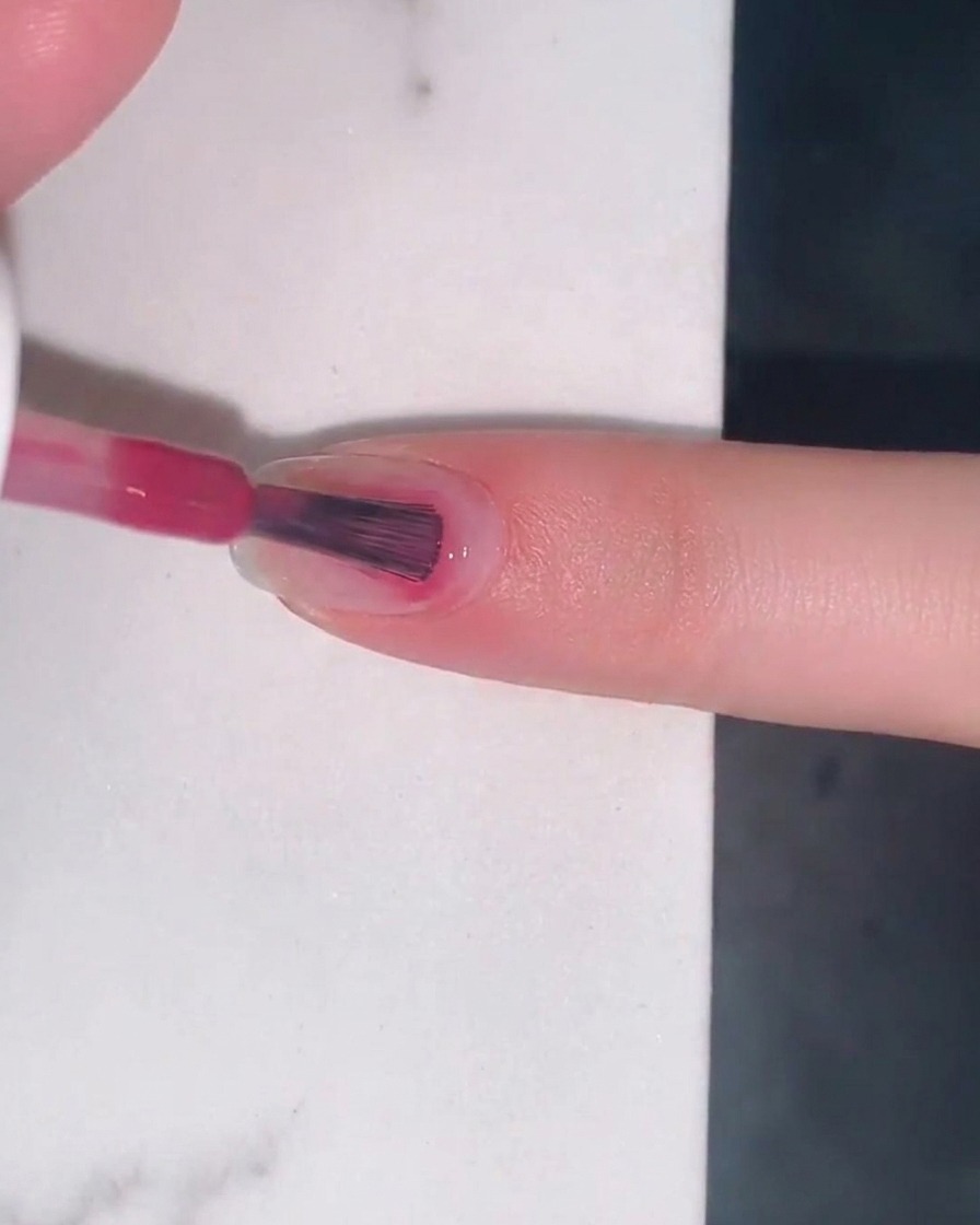 Un pinceau appliquant une fine couche de vernis gel jelly rose baie sur un ongle naturel.