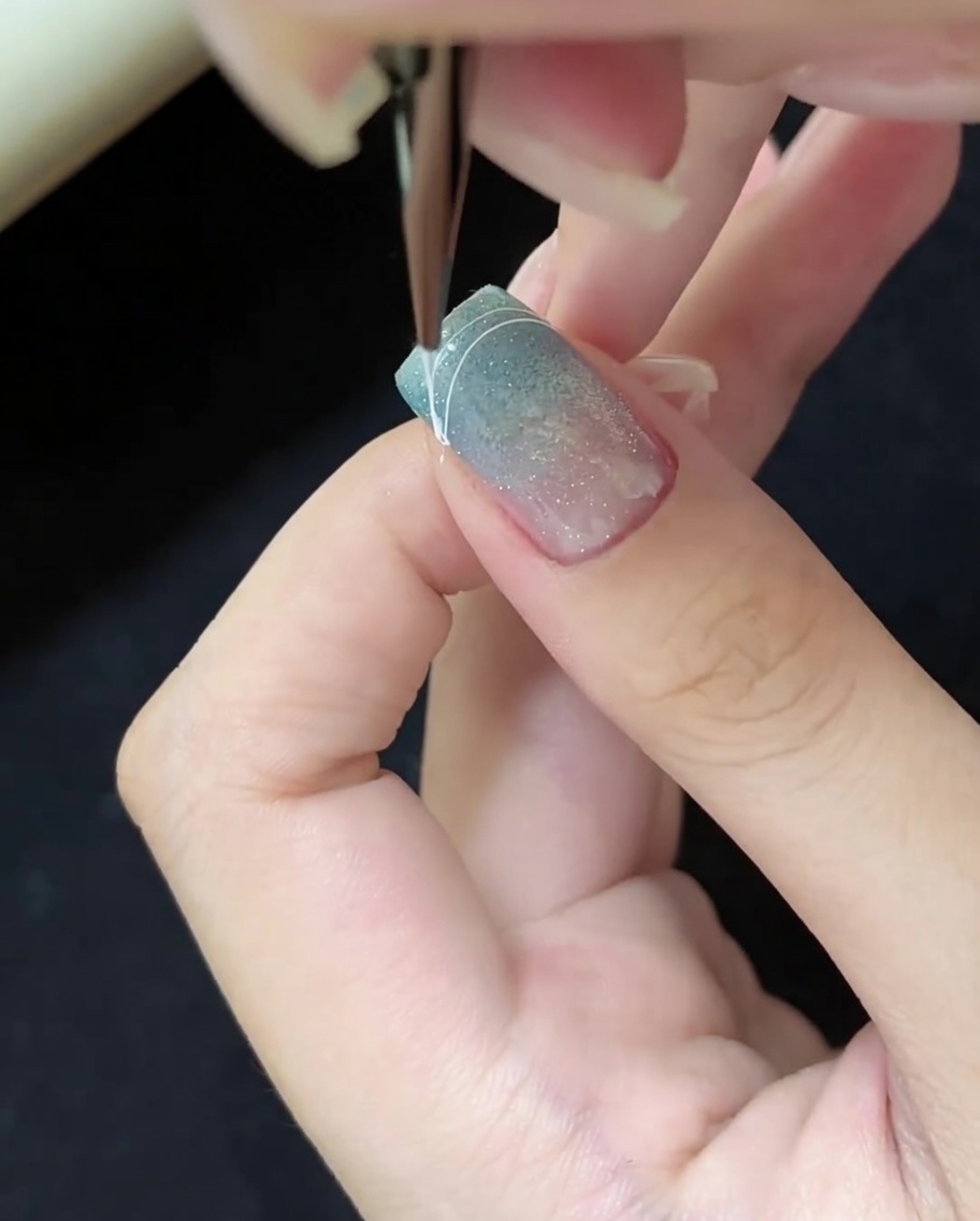 Gros plan dun pinceau dessinant une ligne incurvée blanche sur le coin droit dun ongle cat-eye bleu.
