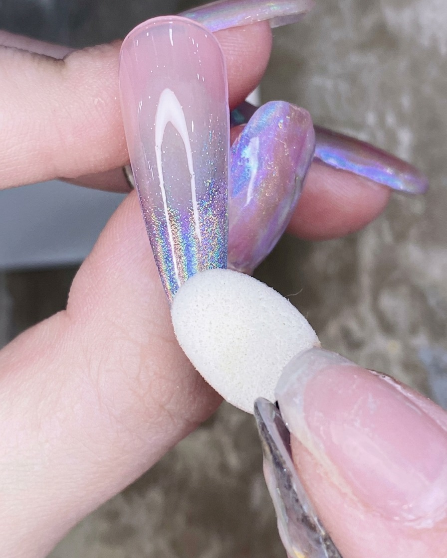 Gros plan dun doigt tenant un ongle avec un applicateur éponge brossant la poudre chrome aurore dans un mouvement de dégradé vers le haut.