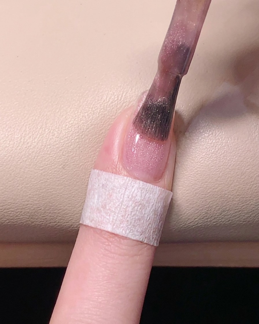 Vue plongeante montrant un pinceau lissant le vernis gel rose sur la plaque de longle pour une finition uniforme.