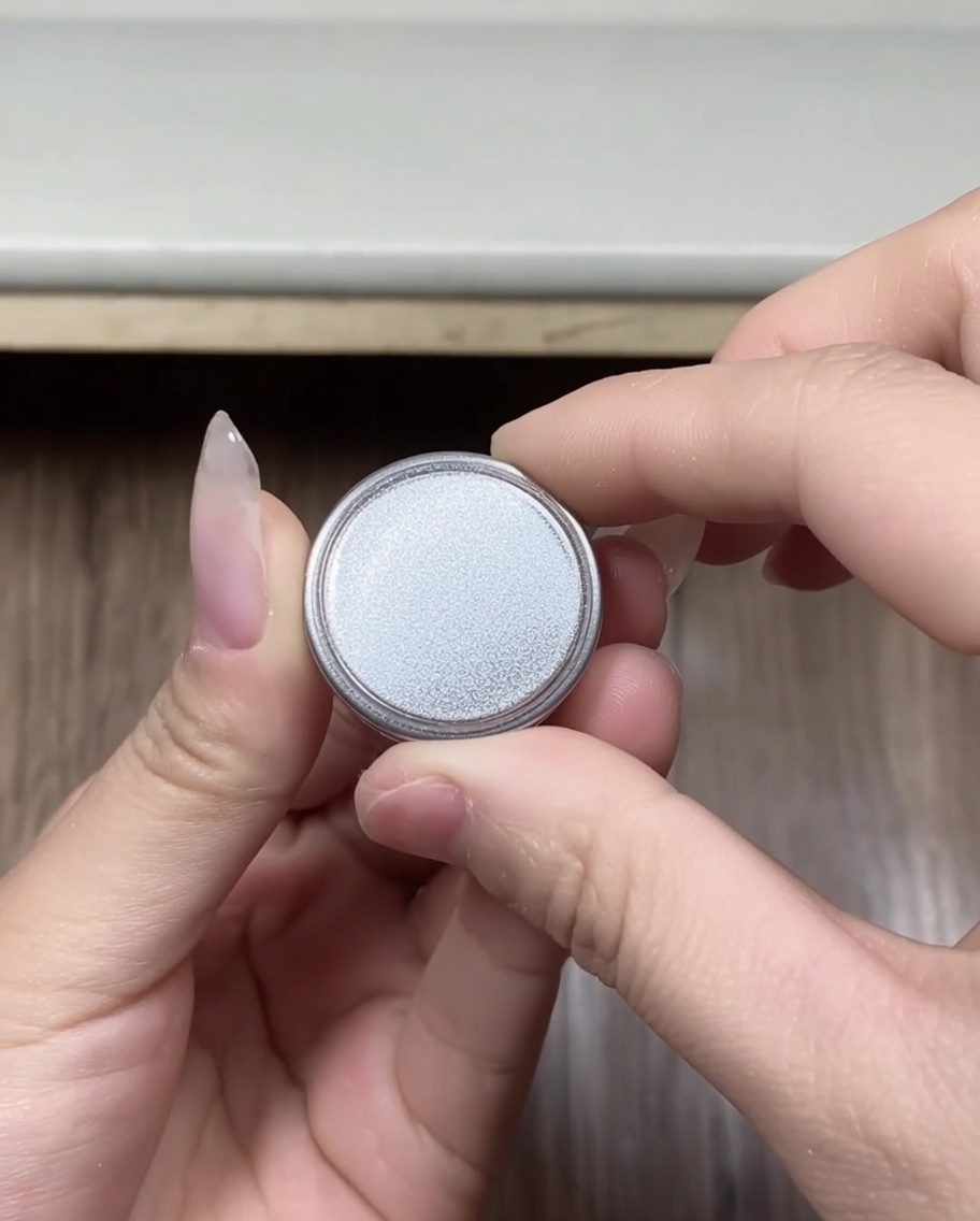 Gros plan de mains tenant un petit récipient rond de poudre chrome argentée, le couvercle juste retiré.