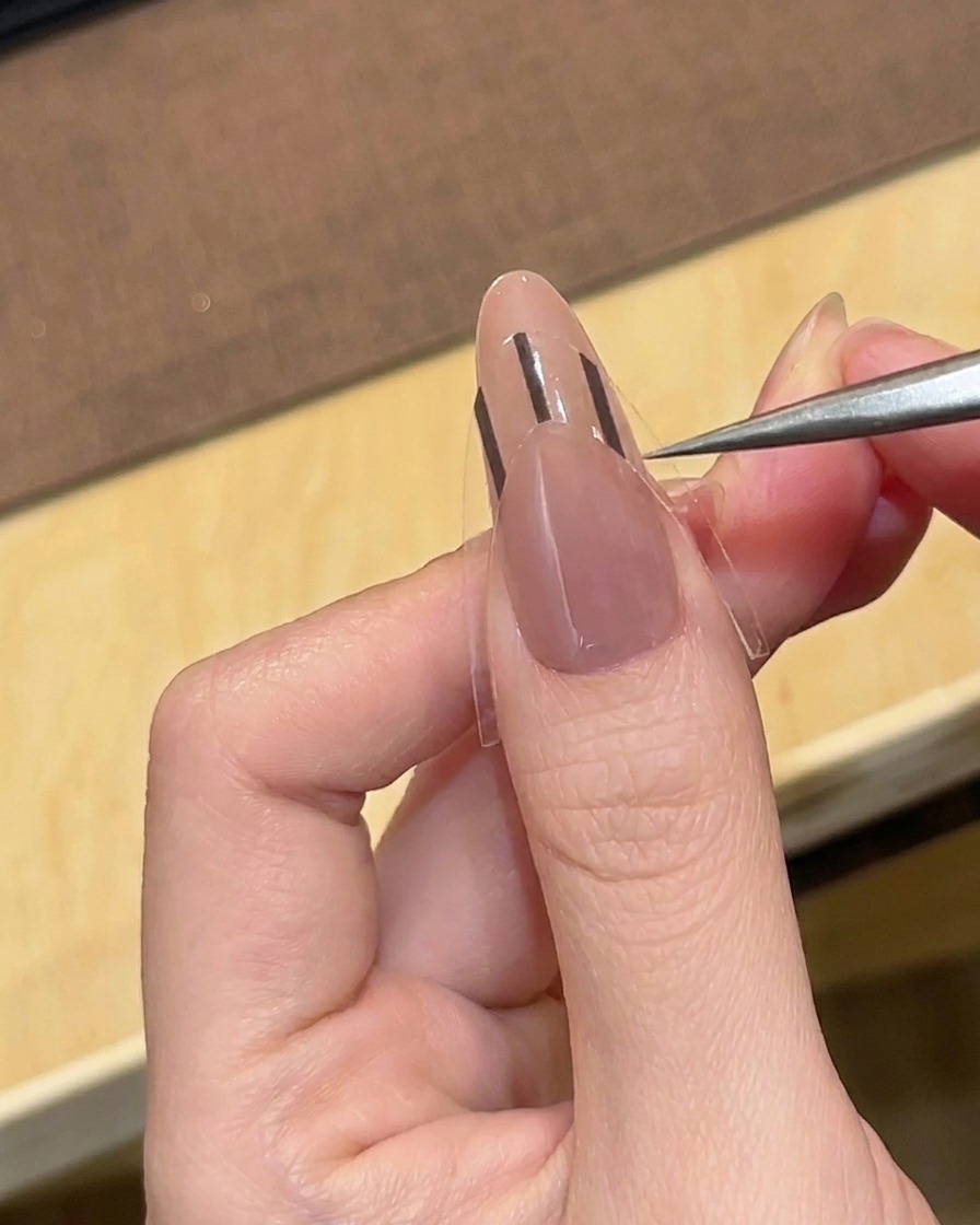 Une pince à épiler plaçant un guide autocollant pour French manucure noir sur un ongle amande de couleur nude.