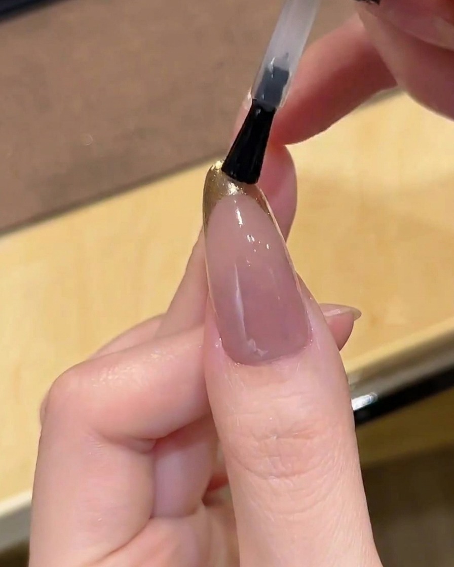 Un pinceau appliquant un top coat transparent et brillant sur un ongle avec une French chromée dorée.