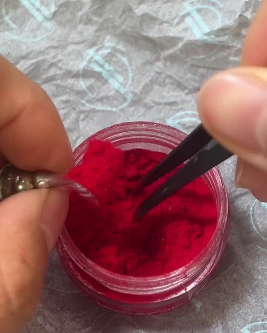 Pinces saupoudrant doucement la poudre rouge sur longle recouvert de gel humide.