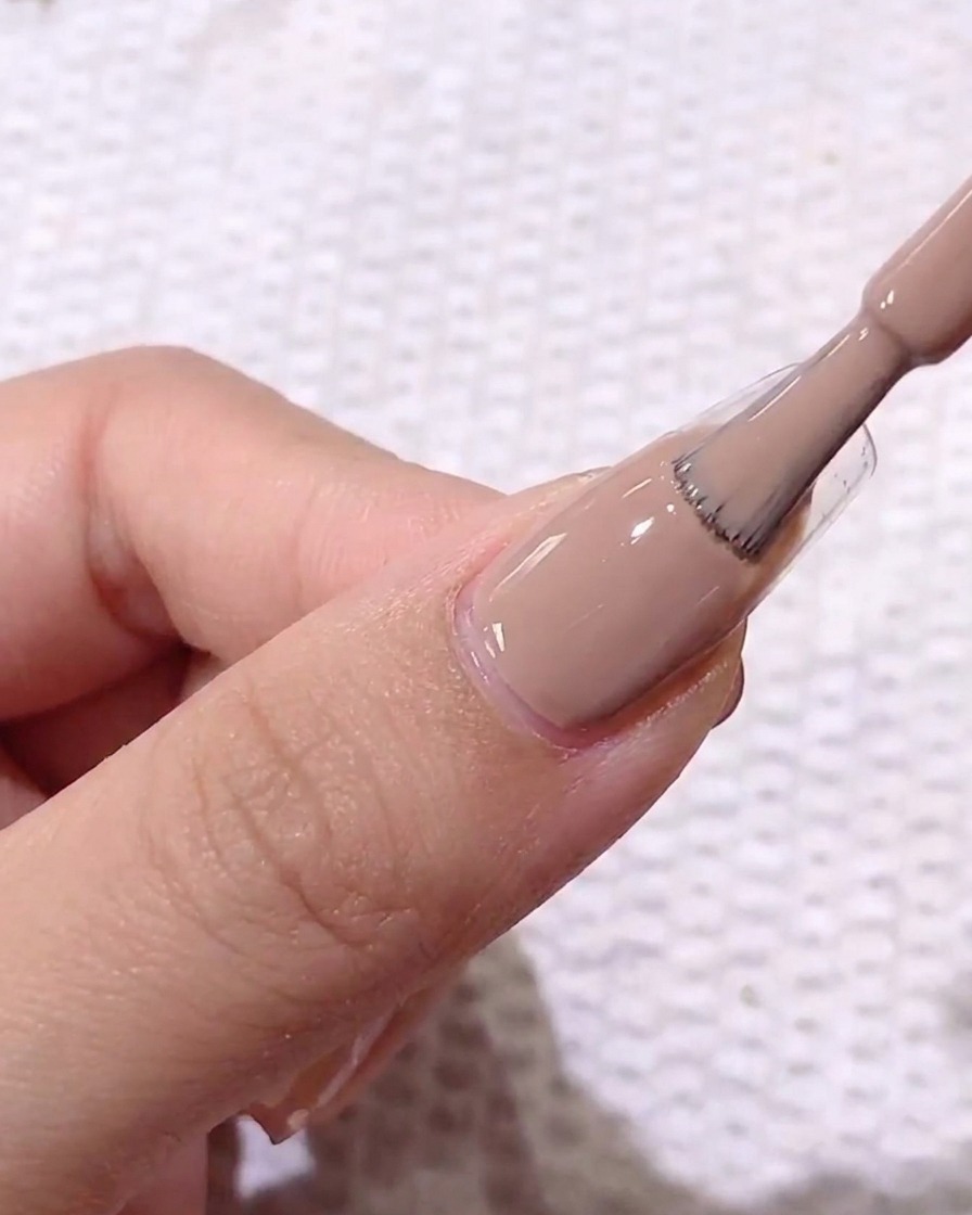 Gros plan sur un pinceau appliquant un vernis gel crème marron clair sur un ongle naturel préparé.