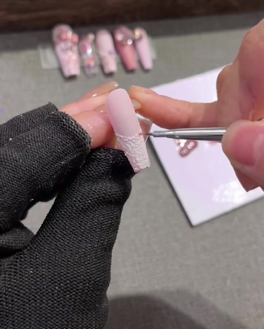 Gros plan dun pinceau liner appliquant de délicates rayures verticales rose clair sur une french tip blanche texturée.