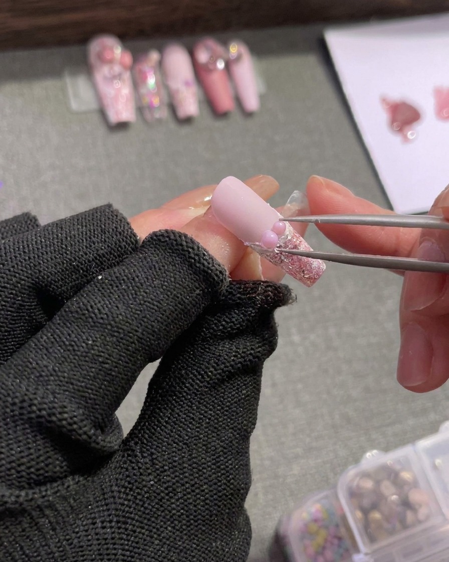Une pince positionnant soigneusement de minuscules strass argentés et une petite breloque en forme de nœud rose à côté dune perle sur la bordure de la french tip.