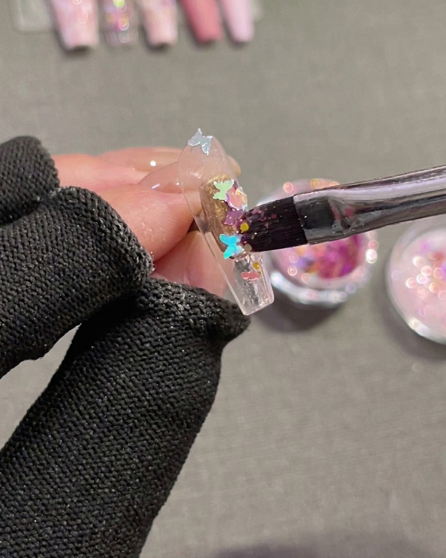 Gros plan dun pinceau utilisé pour presser des sequins holographiques en forme de papillon sur une capsule dongle transparente recouverte de gel humide non catalysé.
