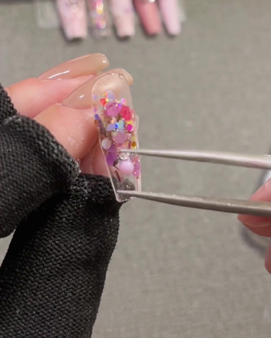 Gros plan dune pince tenant et positionnant une breloque ruban 3D rose sur un ongle recouvert de gel de construction humide.
