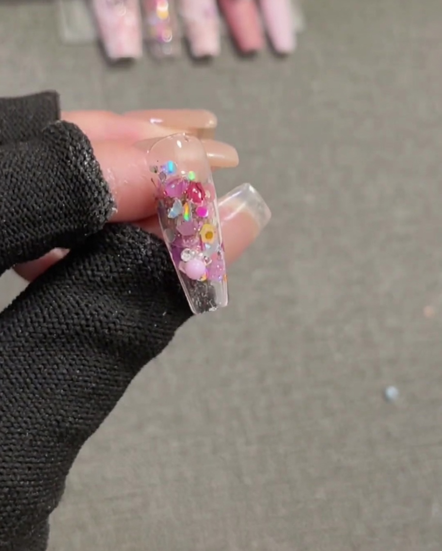 Un pinceau appliquant une dernière couche de top coat transparent ultra-brillant sur un ongle terminé, orné de sequins intégrés et dune breloque nœud rose 3D.