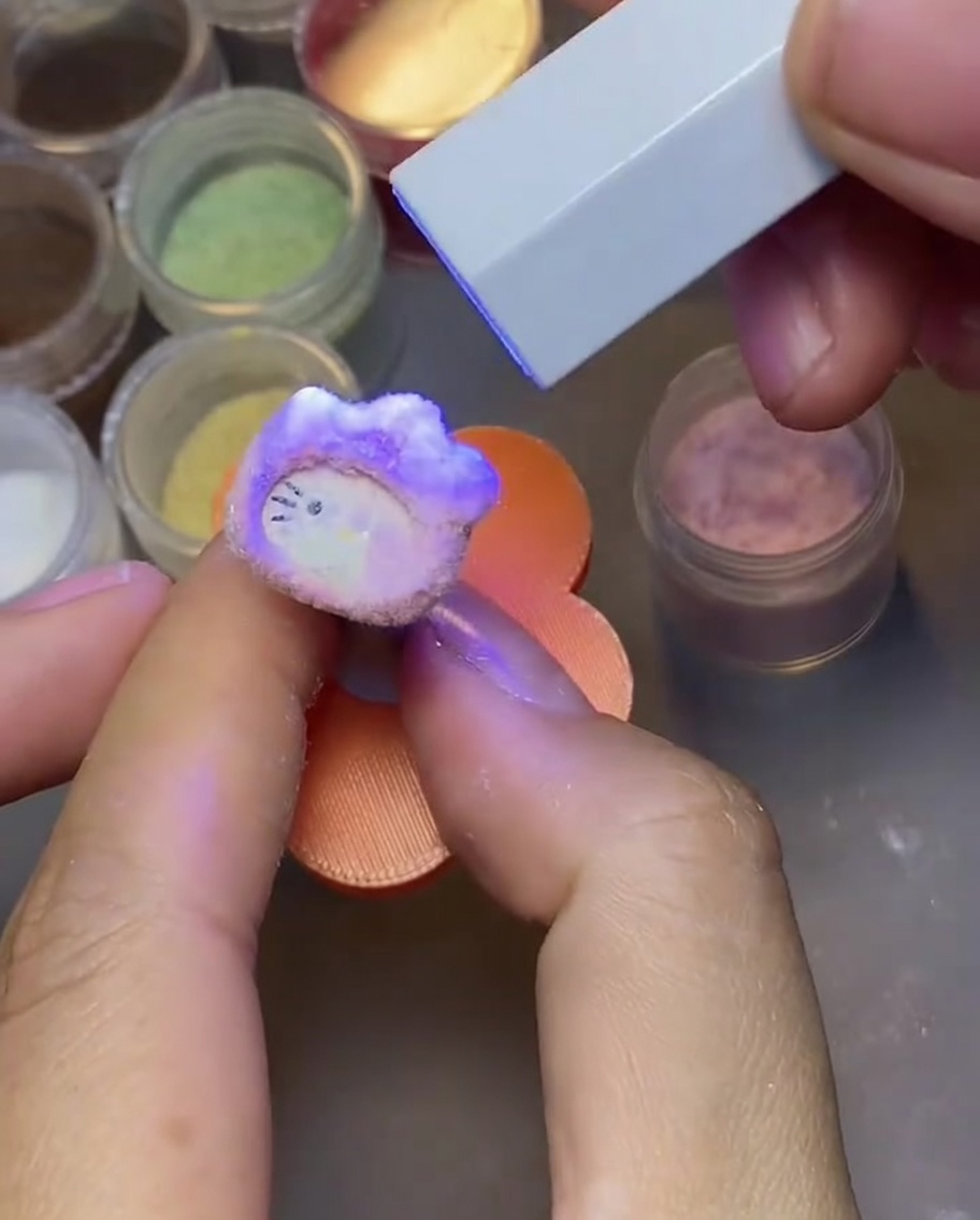 Une main tapote un ongle Hello Kitty catalysé pour enlever lexcédent de poudre velours.