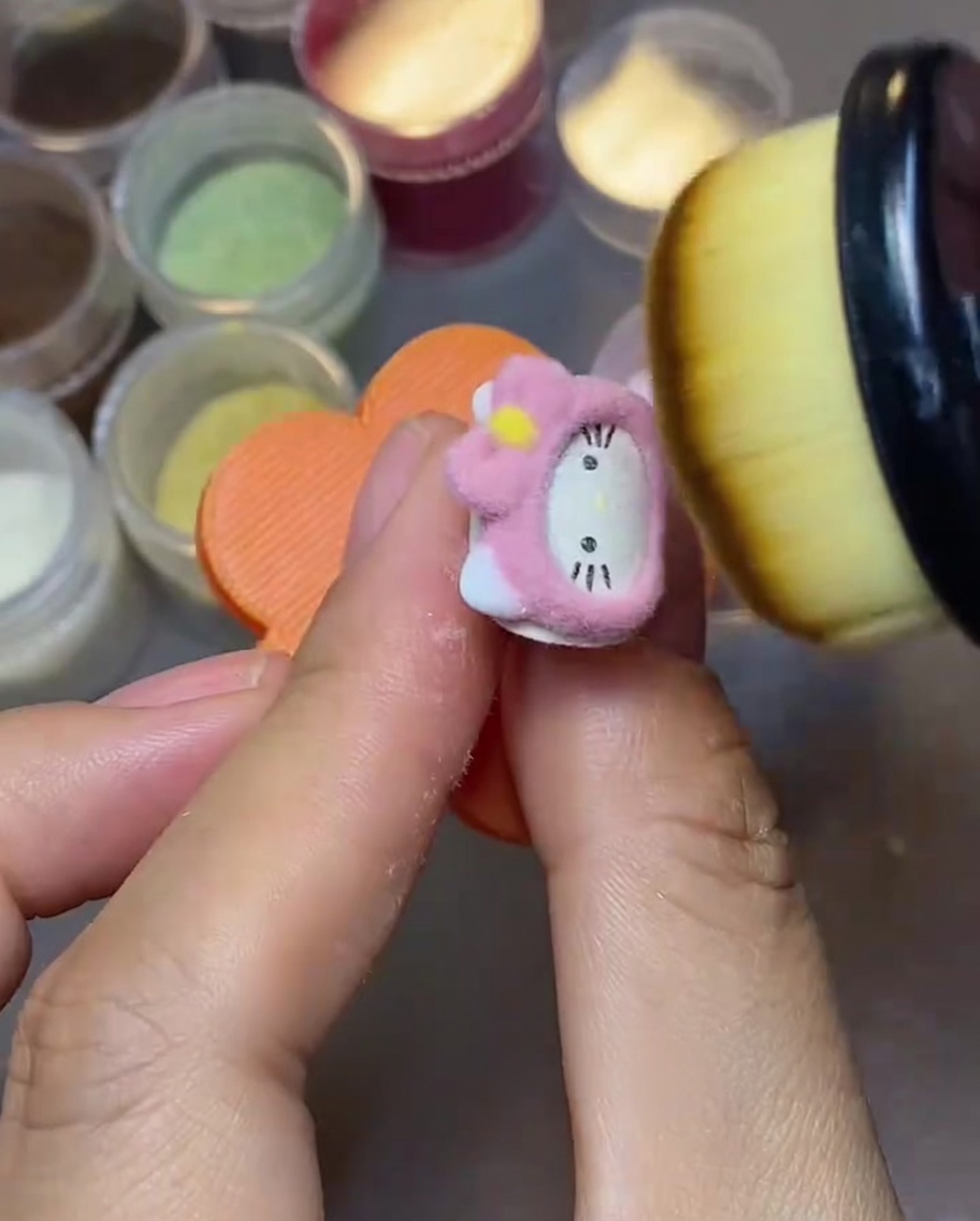 Gros plan dun pinceau à maquillage touffu balayant la poudre velours rose libre sur un ongle Hello Kitty.