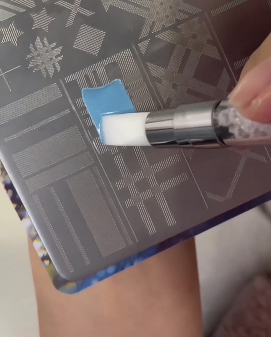 Un pinceau appliquant du vernis bleu clair sur le motif plaid dune plaque de stamping en métal.