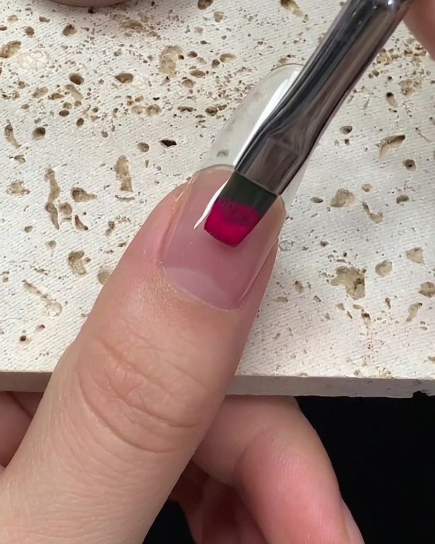 Gros plan sur un pinceau appliquant du vernis gel magenta vibrant sur un ongle, en partant de la cuticule.