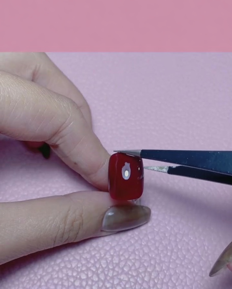 Une main utilisant une pince pour appliquer un morceau de feuille dor sur un ongle rouge brillant.