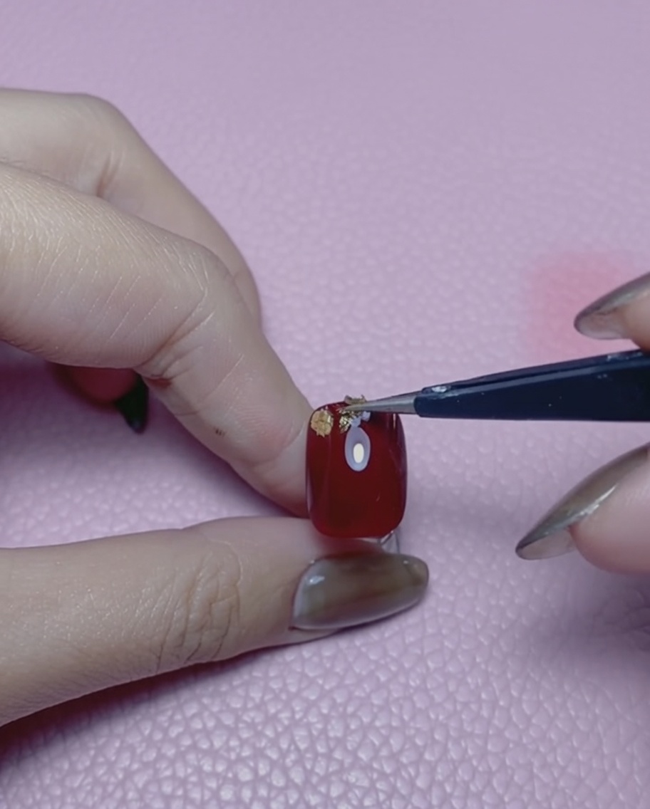 Une paire de pinces tenant un petit morceau de feuille dor au-dessus dun ongle rouge.