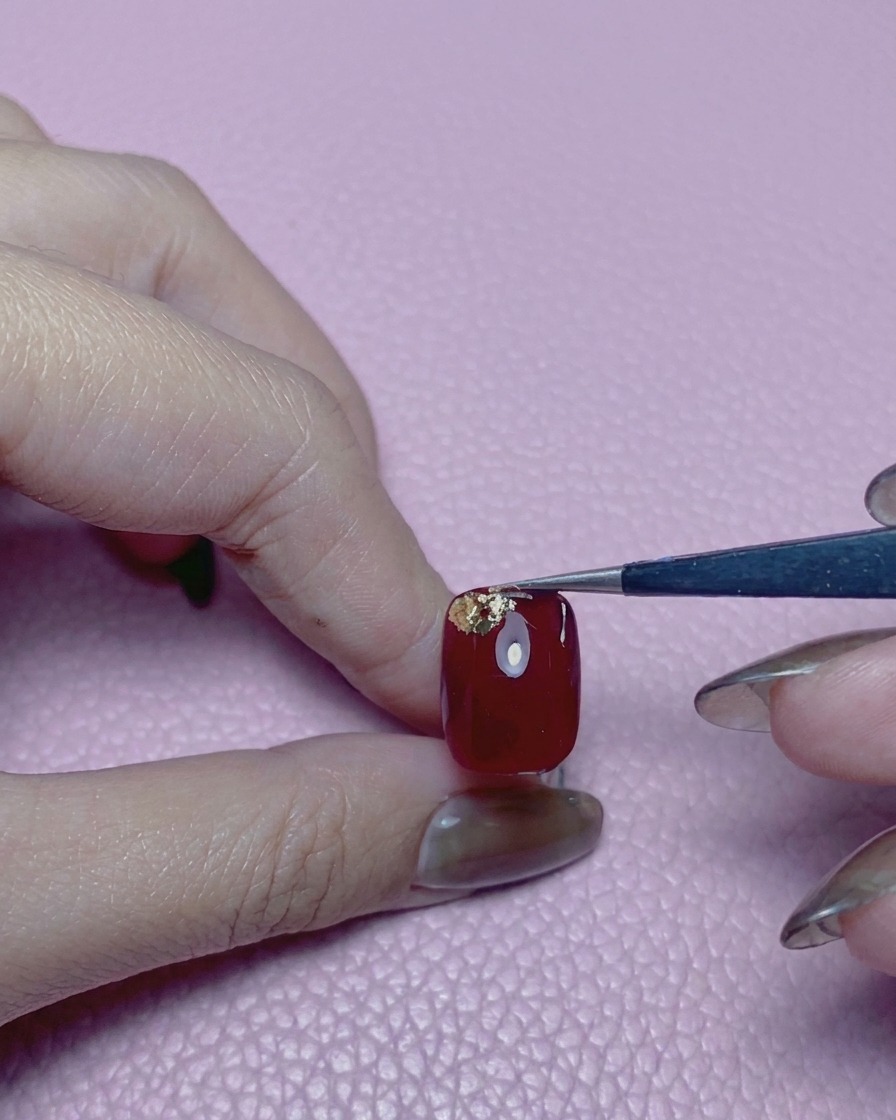 Une pince déposant un troisième morceau de feuille dor sur un ongle rouge vif.