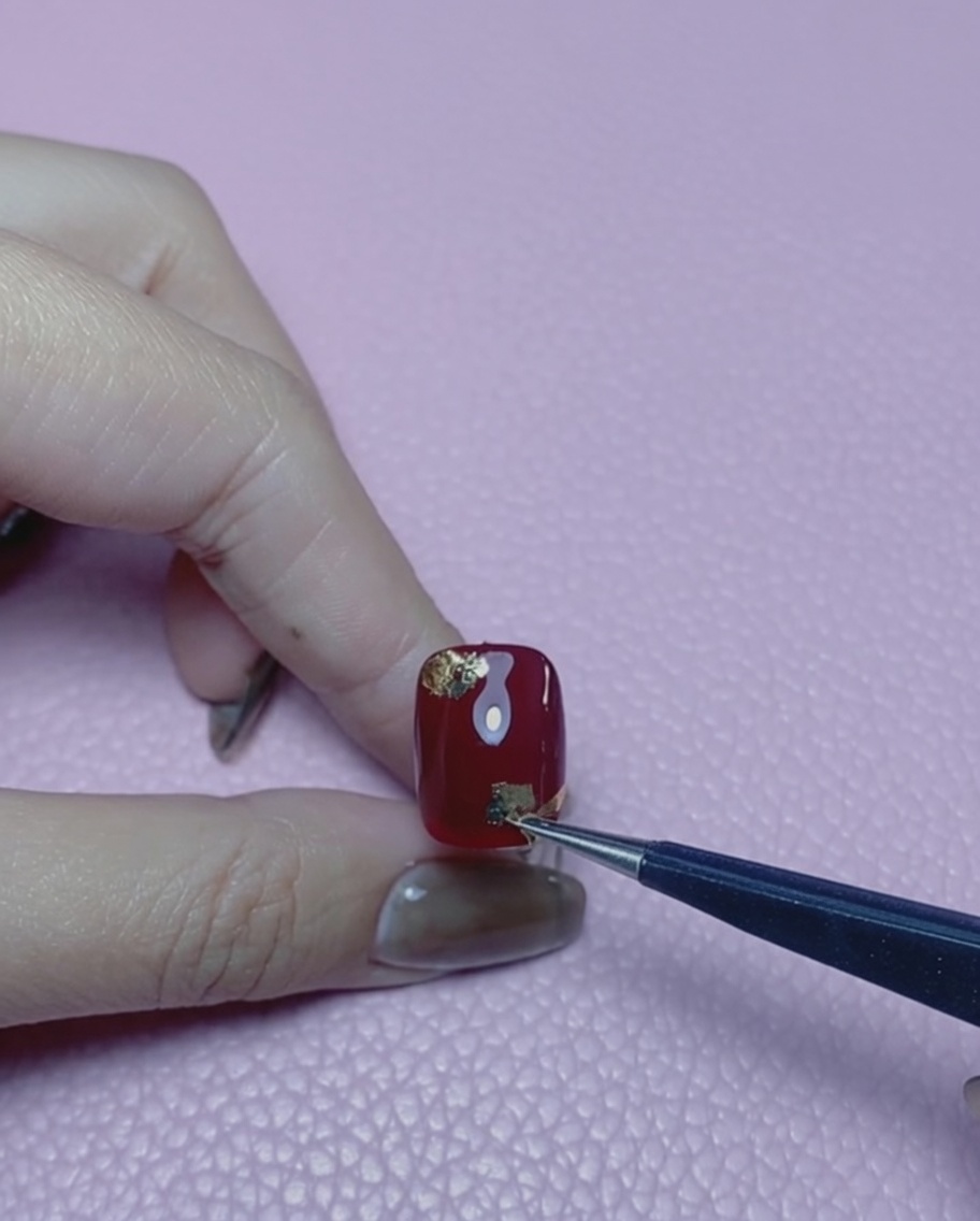 Un outil à embout en silicone pressant les flocons de feuille dor à plat sur un ongle rouge.