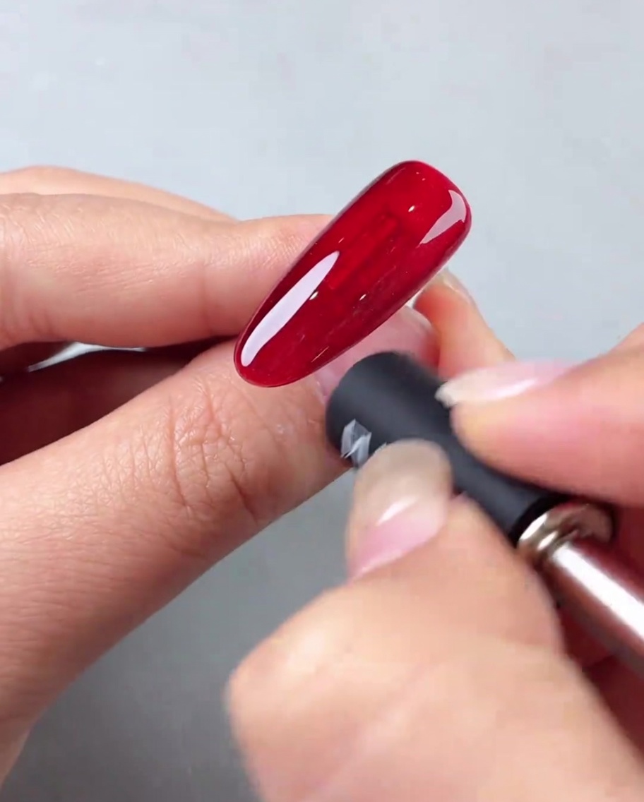 Un aimant à ongles tenu en biais contre un ongle en gel œil de chat rouge pour créer un effet de lumière diagonal.