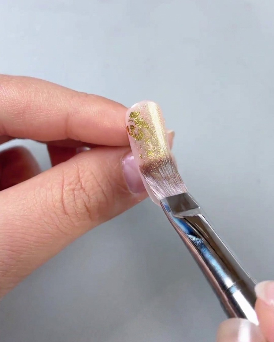 Gros plan dun pinceau appliquant un gel doré scintillant sur des morceaux de feuille dor sur la surface dun ongle.