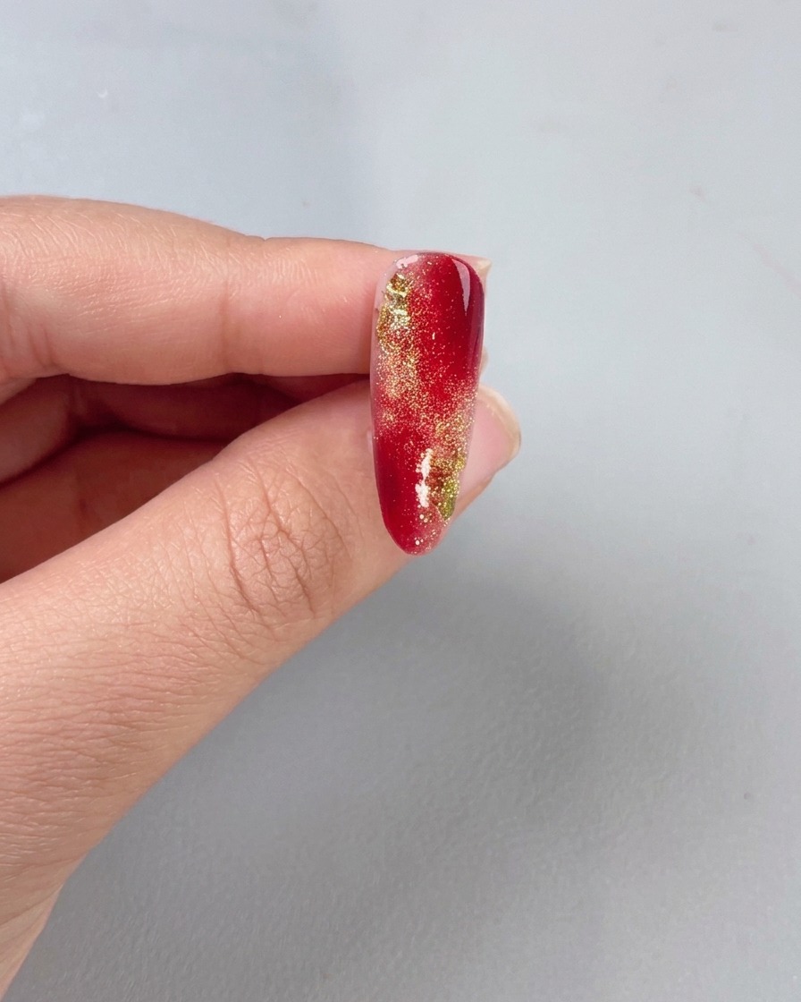 Un pinceau appliquant une couche de finition transparente sur un ongle dégradé rouge et or.