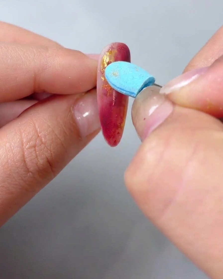 Gros plan dune main utilisant un applicateur éponge bleu pour frotter de la poudre dor métallique sur les lignes texturées en relief dun ongle marbré rouge.