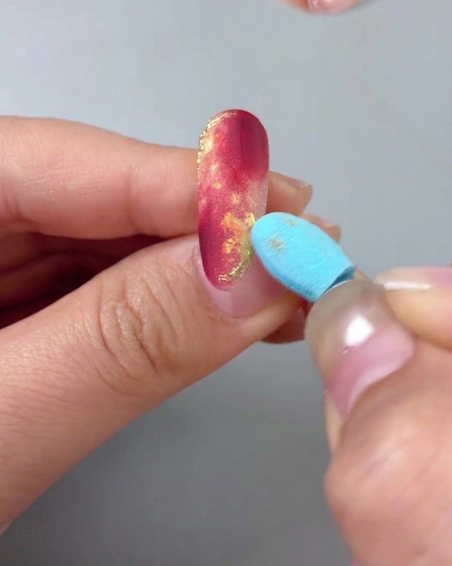 Gros plan dune main avec un ongle marbré rouge et or, tandis quun outil éponge bleu polit doucement la surface pour éliminer lexcès de poudre dor.