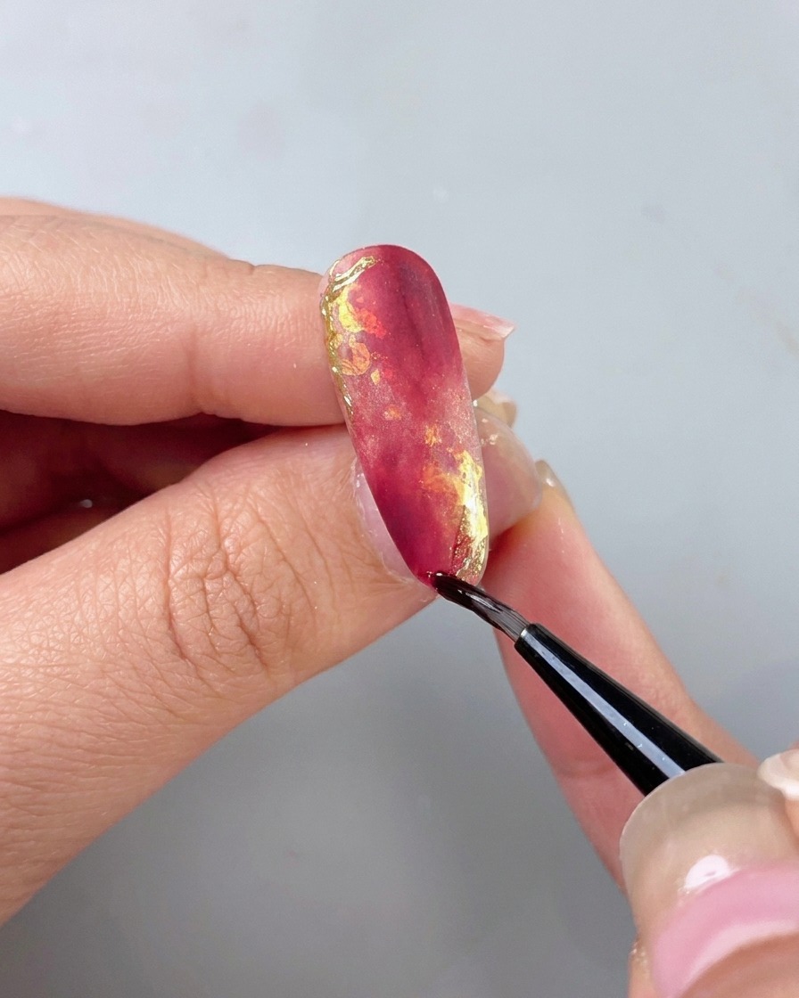 Un pinceau fin appliquant une couche de finition isolante transparente sur un ongle marbré rouge et or magnifiquement décoré.