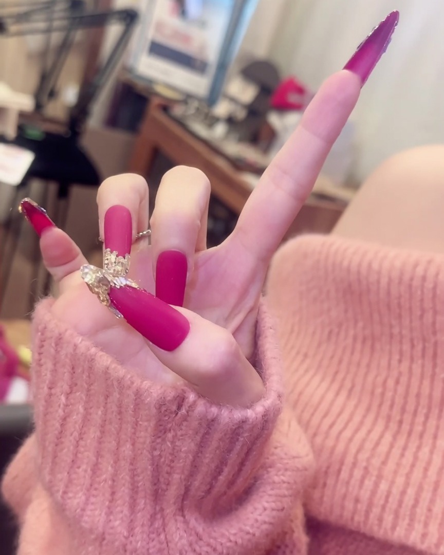 Main aux doigts écartés, montrant les longs ongles rose mat avec accents de cristaux accompagnés de bagues argentées élégantes.